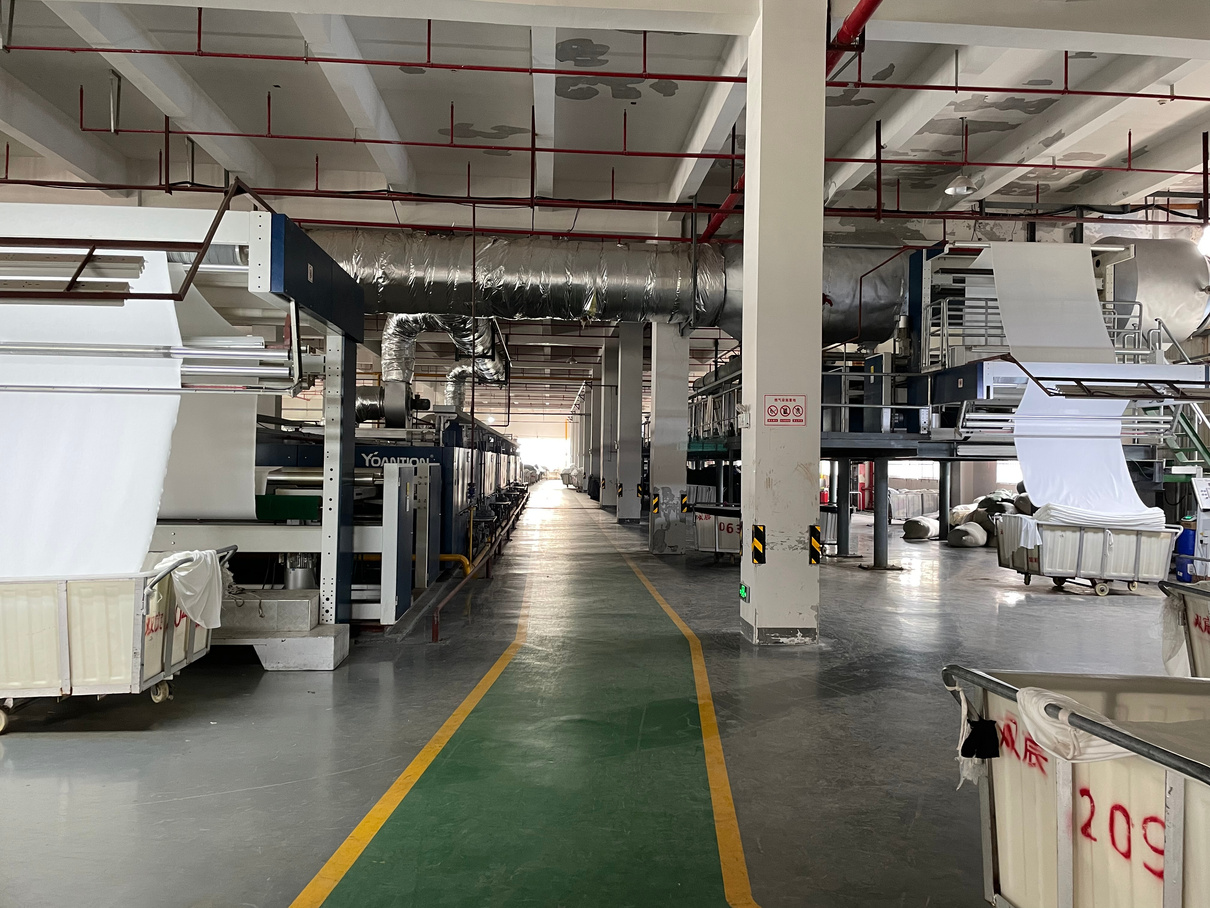 Dye house finishing line in Changshu: fabric feeding through a stenter, trolleys of greige knit along a central aisle.